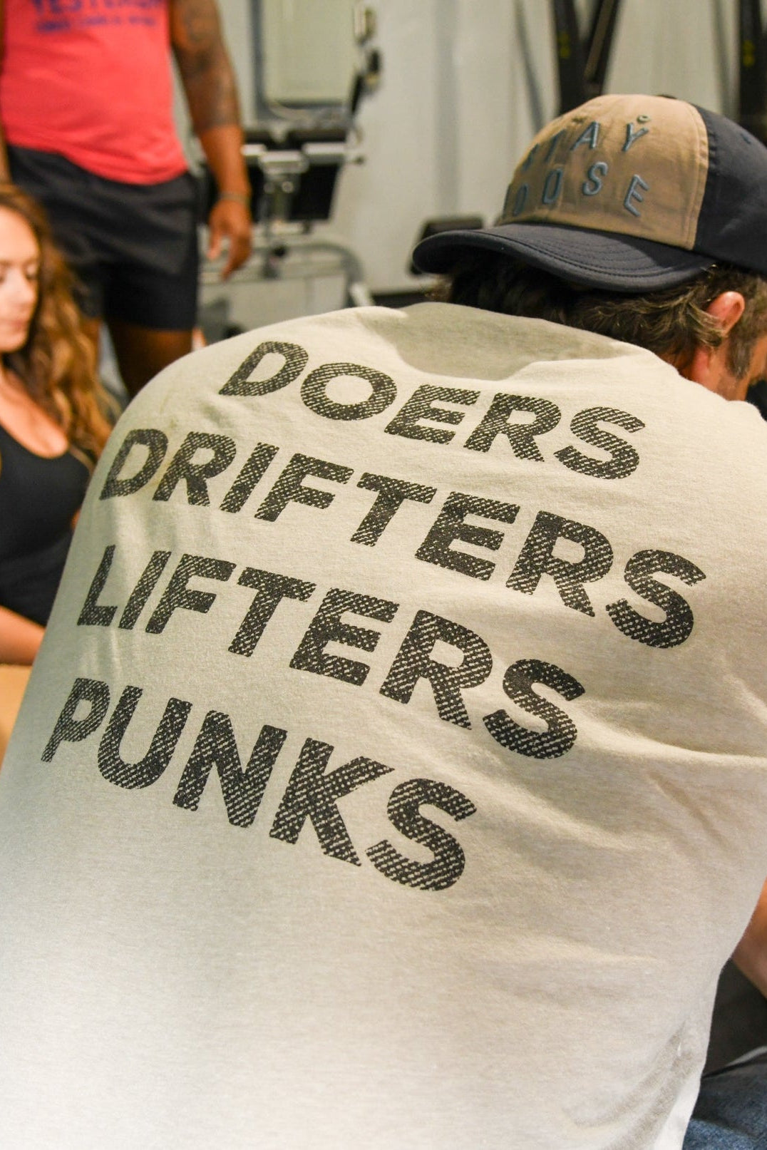 A man in fitness gear wears the Tango Charlie Apparel Doers Drifters Lifters Punks Unisex Tee while sitting indoors with two women; gym equipment is visible. Show off your crew and style at the gym with this bold tee.