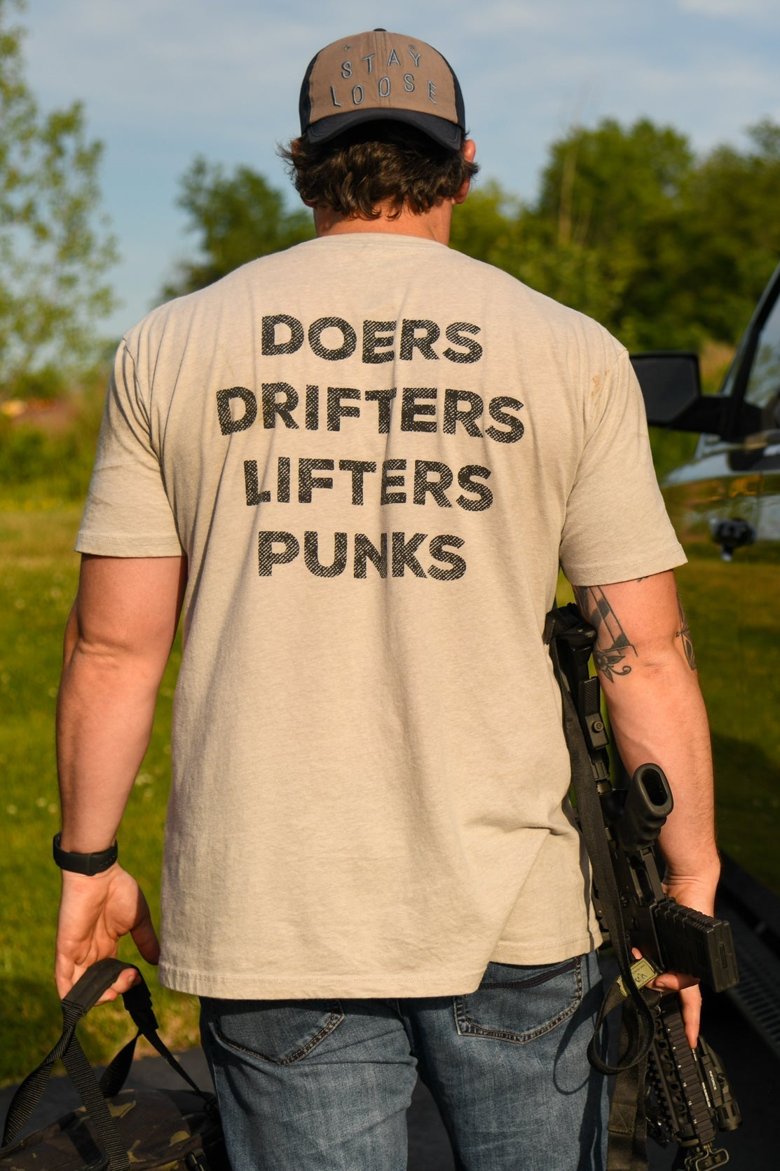 Wearing the Doers Drifters Lifters Punks Unisex Tee by Tango Charlie Apparel, a person stands outdoors by a black SUV with a rifle and bag. Trees and grass frame the scene—fitness gear for those who rep their crew boldly.