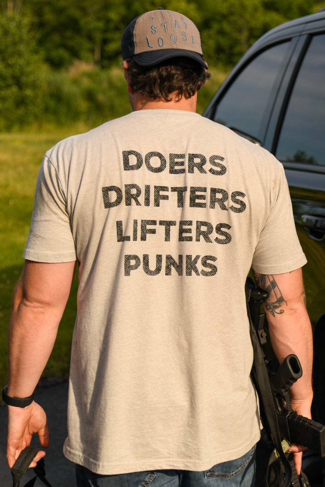 A man wears the Doers Drifters Lifters Punks Unisex Tee by Tango Charlie Apparel, paired with a cap and beige fitness wear, standing outdoors by a black SUV with a rifle—rep your crew in style.