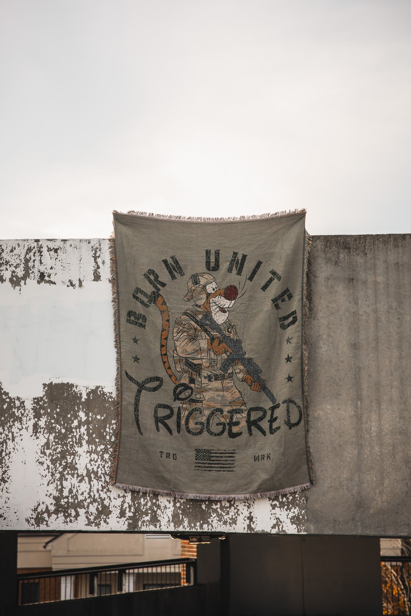 The Triggered Jacquard Blanket by Born United Private Label features a bold tiger in military gear with a rifle against a concrete wall, surrounded by stars and flags, and includes the text "Born United Triggered." Fringed edges complete the look.