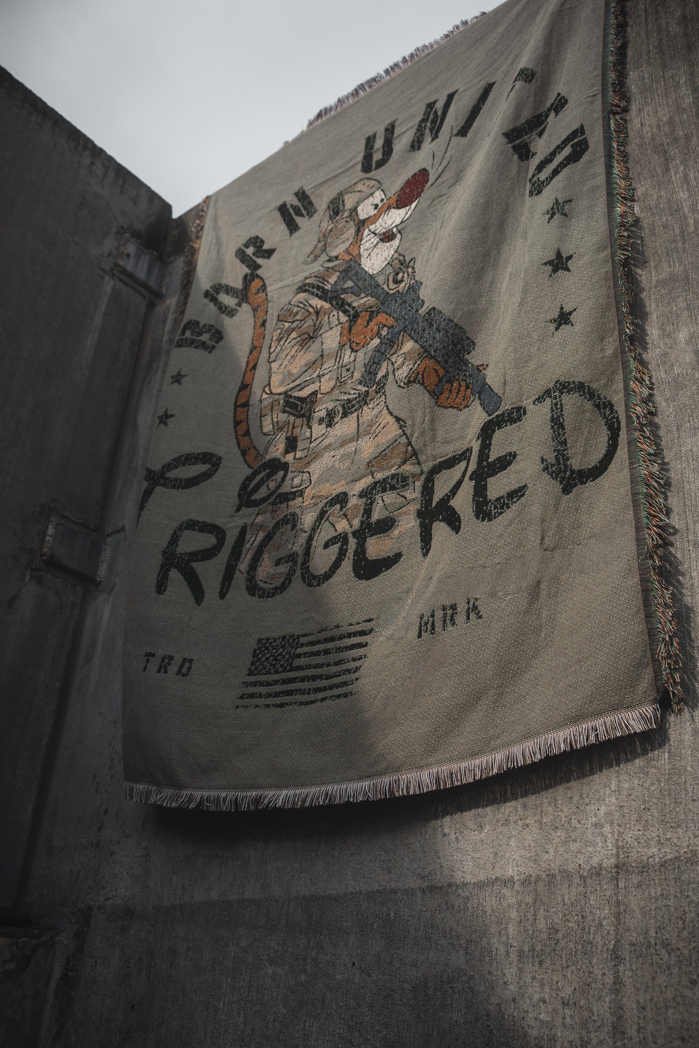 The Triggered Jacquard Blanket by Born United Private Label hangs against a concrete wall, showcasing a tiger in tactical gear with a rifle and bold “BURN UNIT” and “TRIGGERED” lettering on its fringed edges.