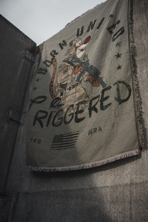 The Triggered Jacquard Blanket by Born United Private Label hangs against a concrete wall, showcasing a tiger in tactical gear with a rifle and bold “BURN UNIT” and “TRIGGERED” lettering on its fringed edges.