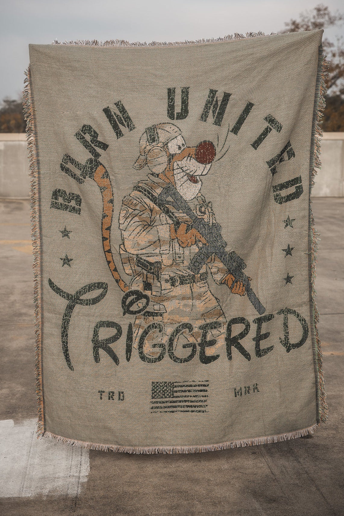 The Triggered Jacquard Blanket by Born United Private Label, featuring an armed soldier in tactical gear with military-style graphics and fringed edges, is displayed outdoors against a concrete background.