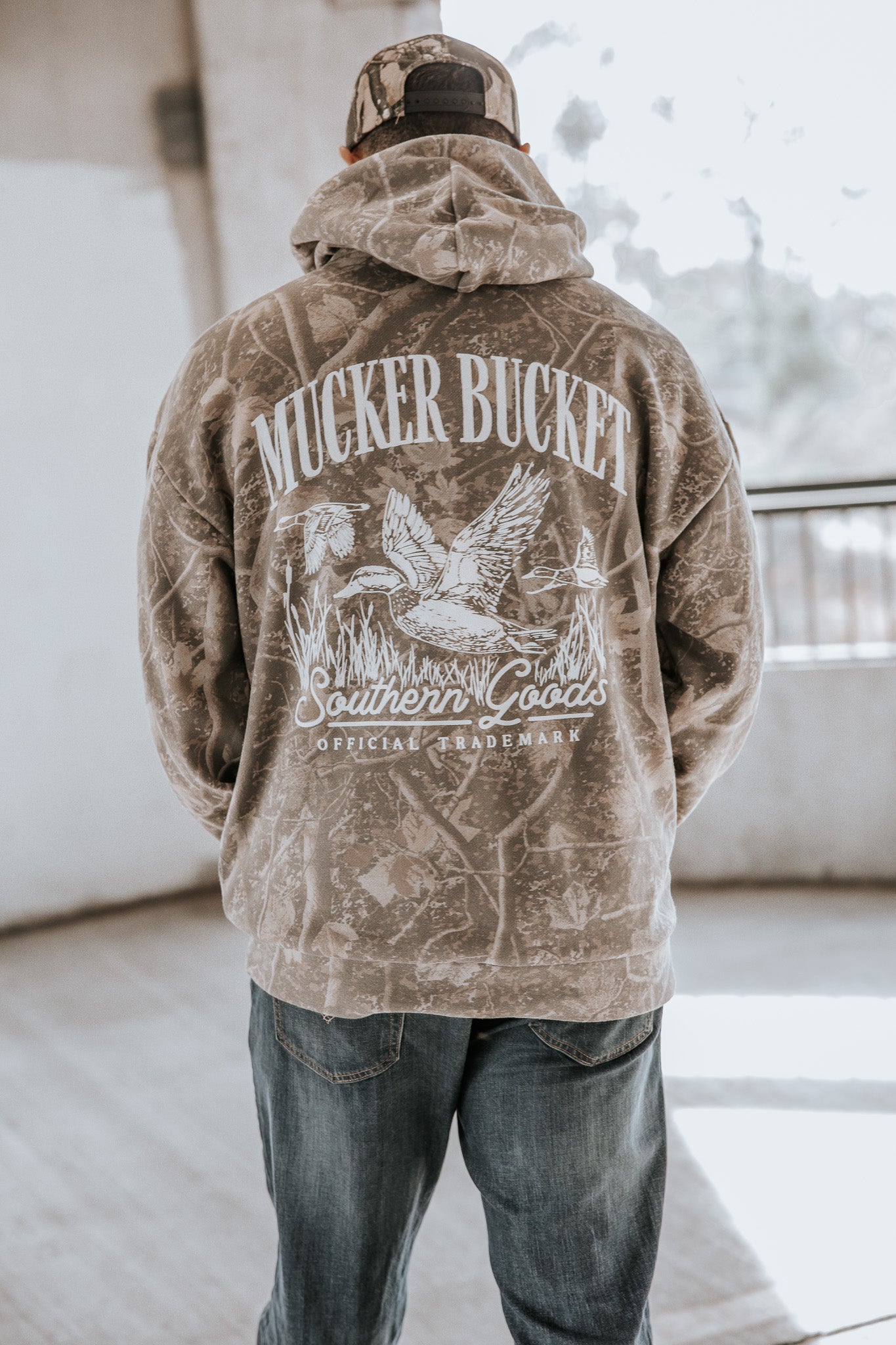 Someone wearing the Mucker Bucket Mallard Deadwood Camo Hoodie featuring the brand’s official trademark and a duck graphic on the back, stands indoors with their back to the camera.