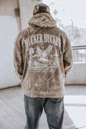 Someone wearing the Mucker Bucket Mallard Deadwood Camo Hoodie featuring the brand’s official trademark and a duck graphic on the back, stands indoors with their back to the camera.