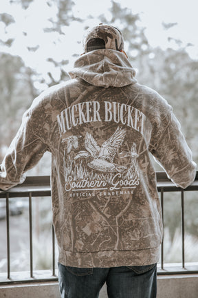 A person in a Mallard Deadwood Camo Hoodie and hat by Mucker Bucket stands at a railing, seen from behind. The hoodie features ducks and reeds along with the brand's name.