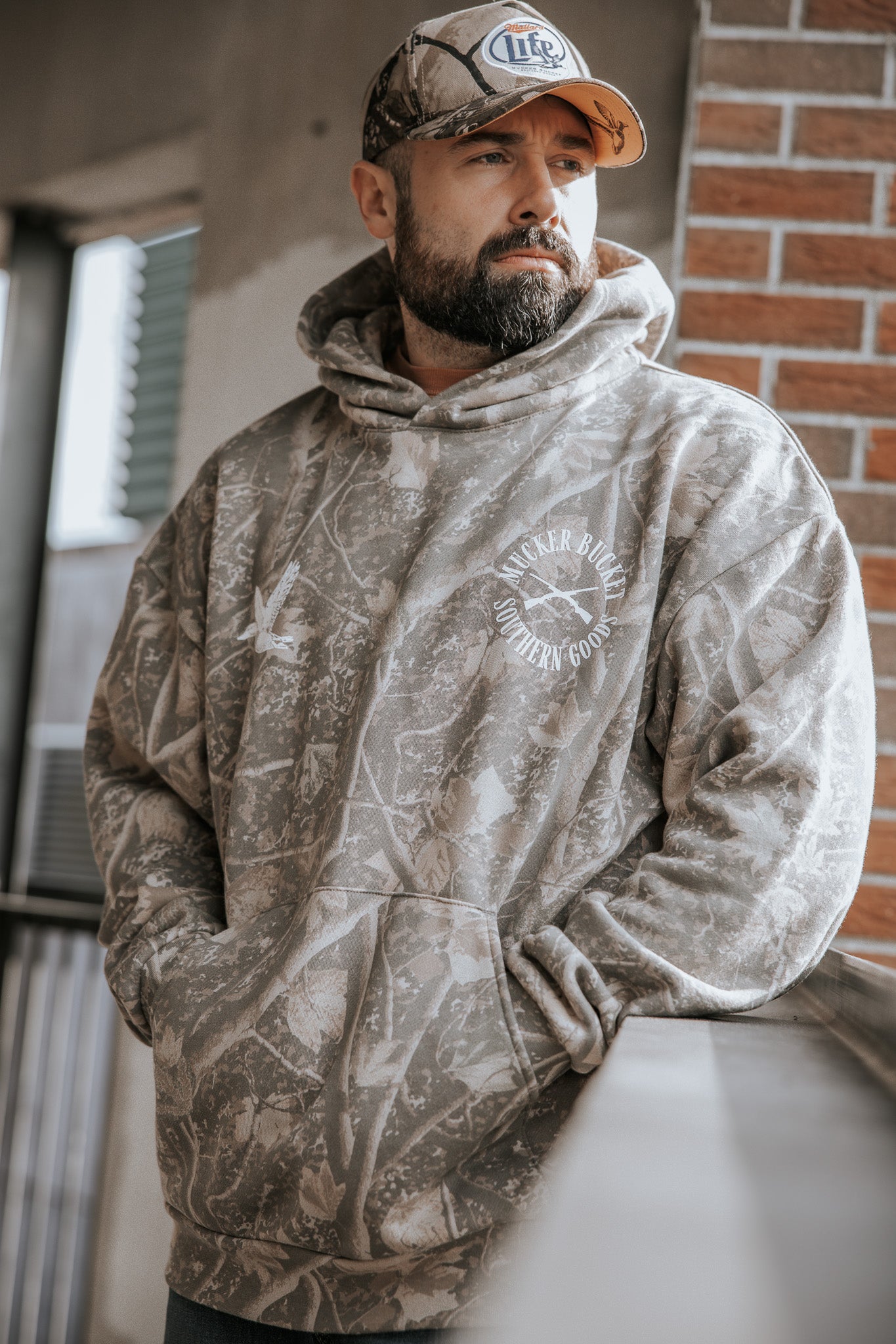 A bearded man in a Mucker Bucket Mallard Deadwood Camo Hoodie and a Miller Lite cap leans on a railing, looking to the side near a brick wall and a partially open window outdoors.