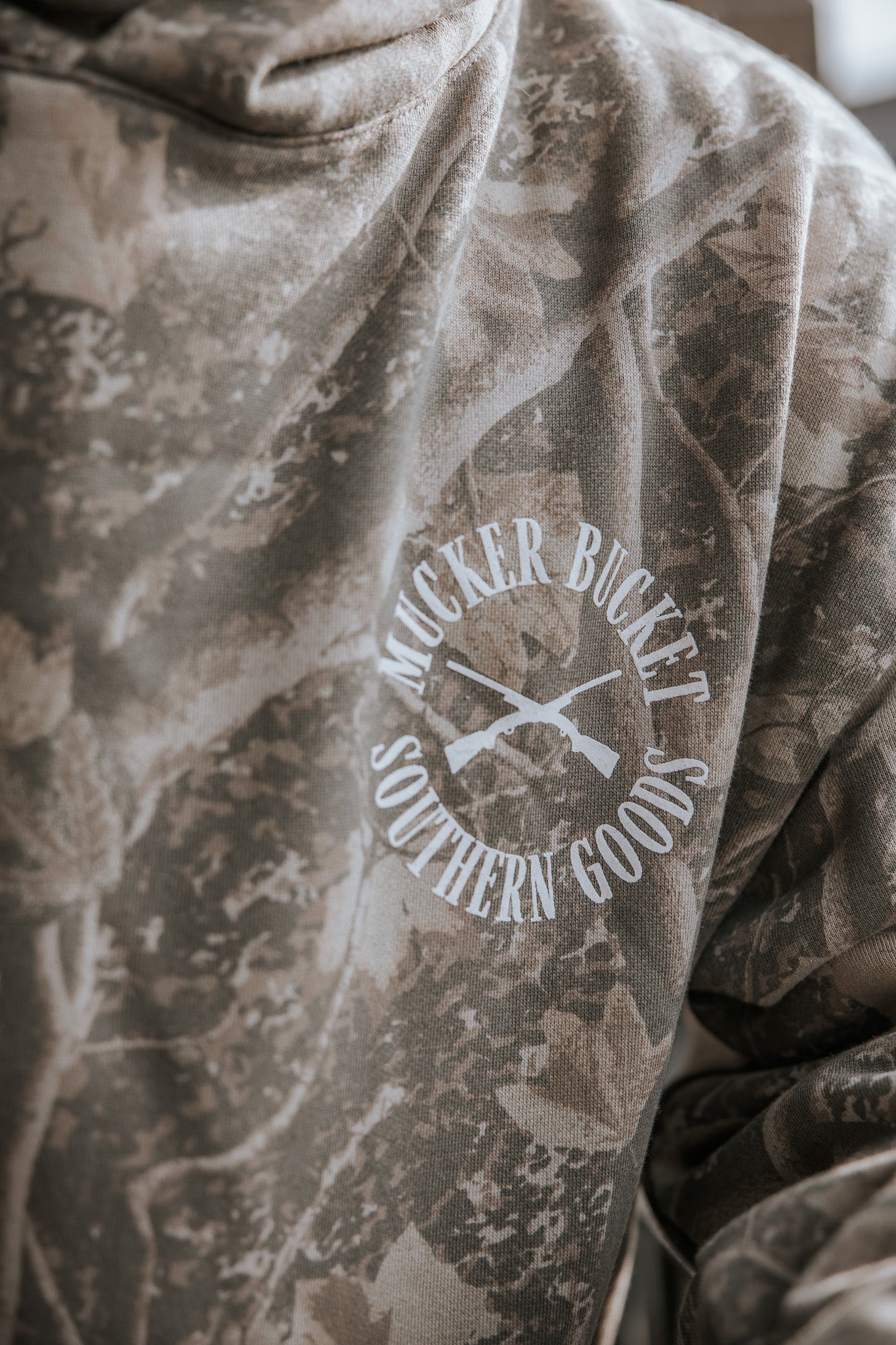 A close-up of the Mallard Deadwood Camo Hoodie by Mucker Bucket, featuring a white logo with two crossed rifles and "Mucker Bucket Southern Goods.