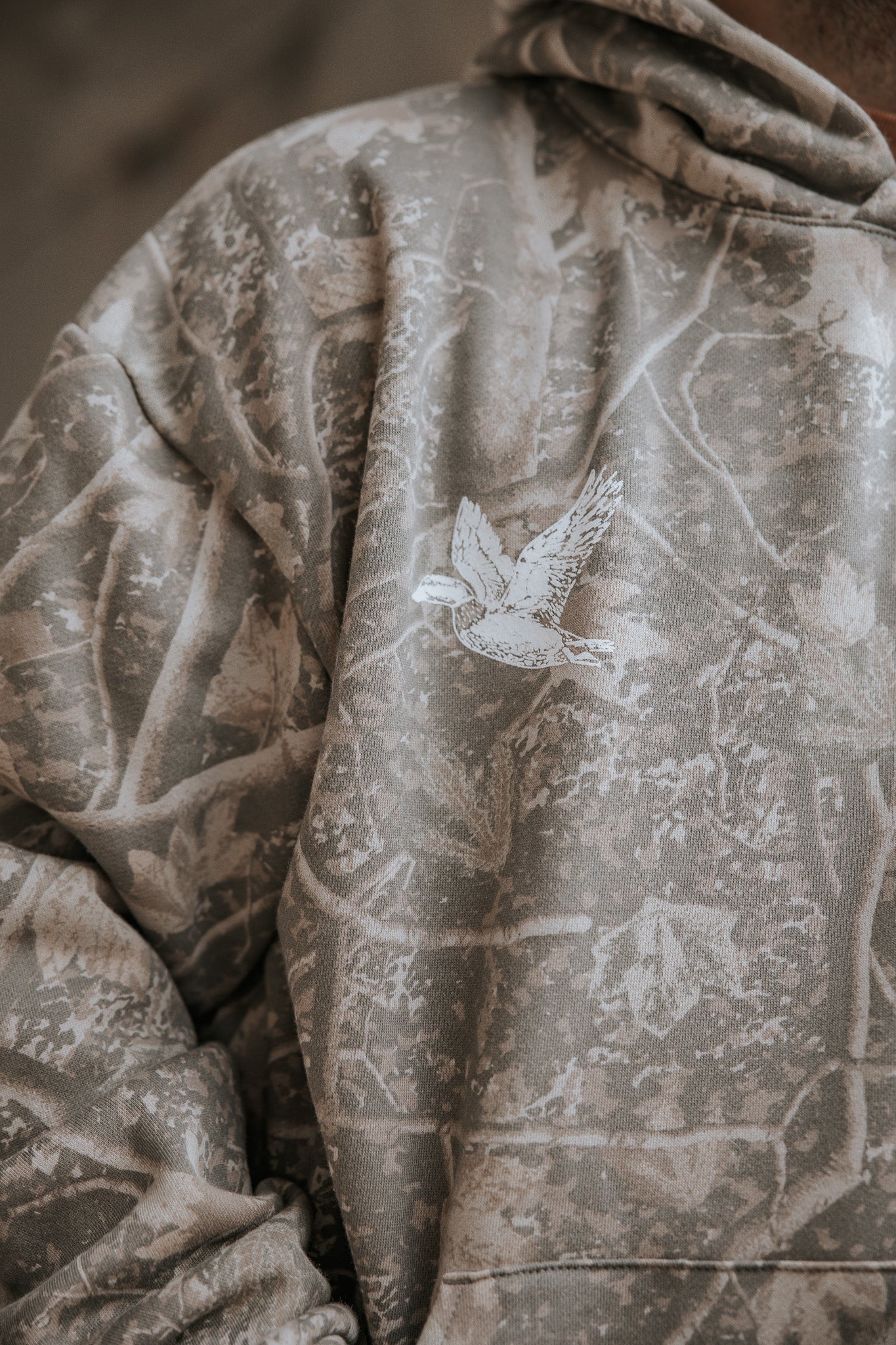 The Mallard Deadwood Camo Hoodie by Mucker Bucket is a premium relaxed fit fleece in gray camouflage, featuring a white bird illustration on the chest and a nature-inspired pattern with branches and leaves.