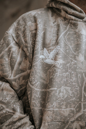 The Mallard Deadwood Camo Hoodie by Mucker Bucket is a premium relaxed fit fleece in gray camouflage, featuring a white bird illustration on the chest and a nature-inspired pattern with branches and leaves.