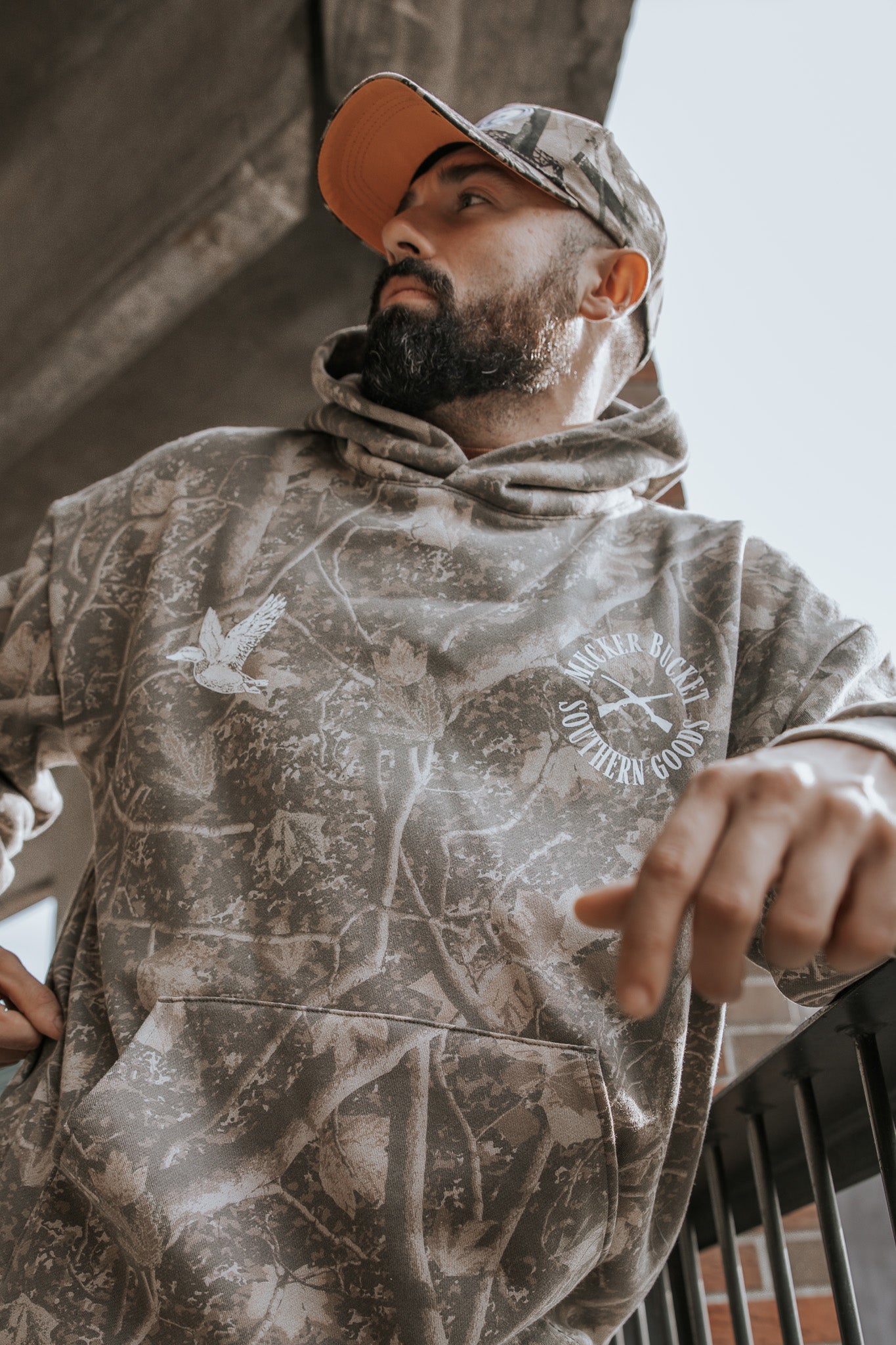 A bearded man in a Mucker Bucket Mallard Deadwood Camo Hoodie and cap leans on a metal railing, looking to the side with a concrete structure visible overhead.