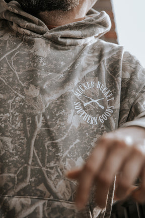 Wearing a Mallard Deadwood Camo Hoodie by Mucker Bucket, featuring a white circle logo with crossed paddles, this person’s face is partly out of frame as they extend their hand forward.