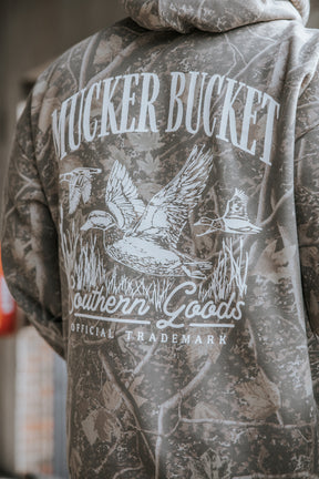 A person wears the Mallard Deadwood Camo Hoodie by Mucker Bucket, featuring the brand’s official trademark and a flying ducks illustration—also known as the stacking ducks hoodie—printed on the back.