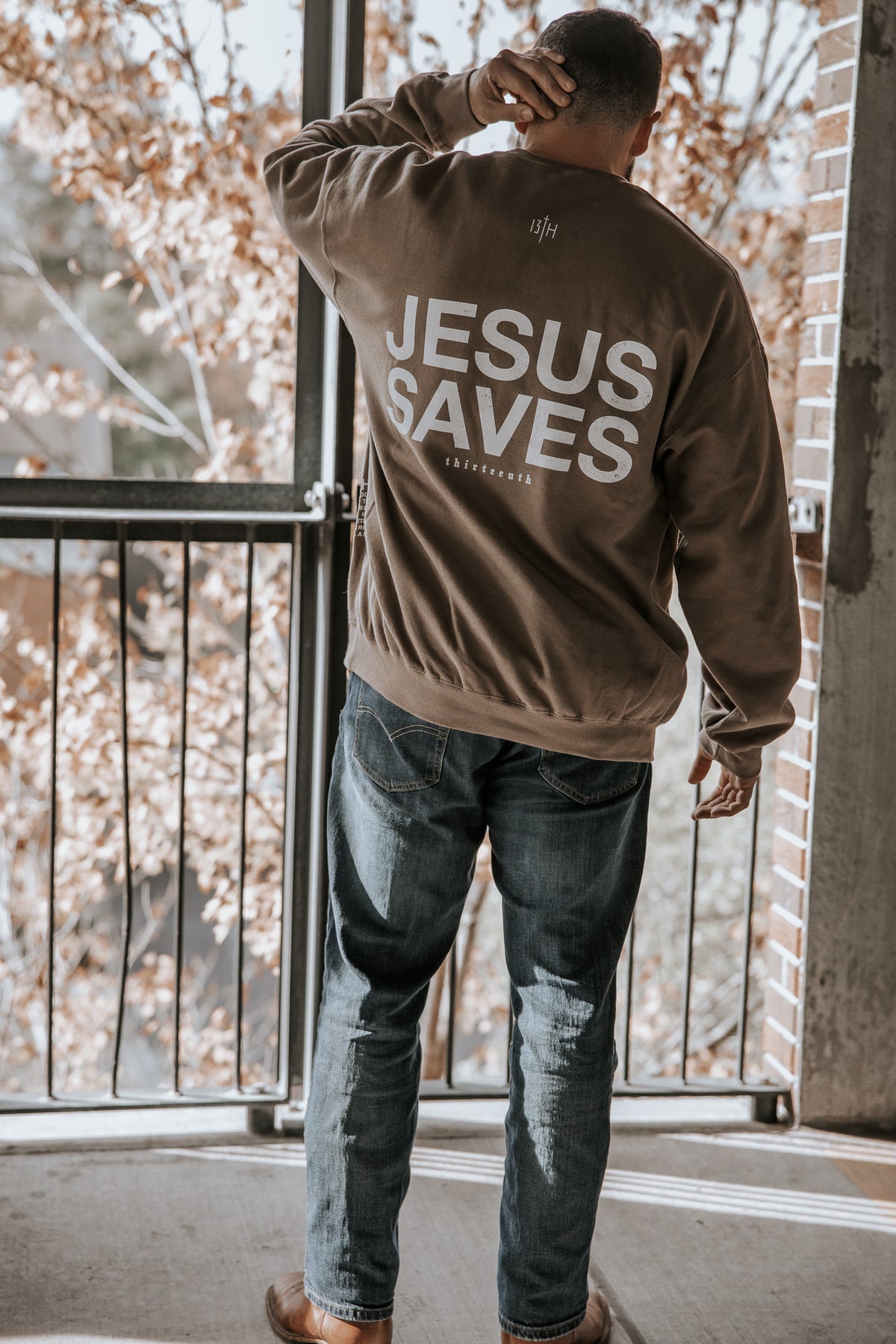 A person stands on a balcony with autumn leaves behind them, wearing the 13th Jesus Saves Crewneck in brown with “JESUS SAVES” printed in large white letters on the back. They are facing away from the camera and scratching their head.
