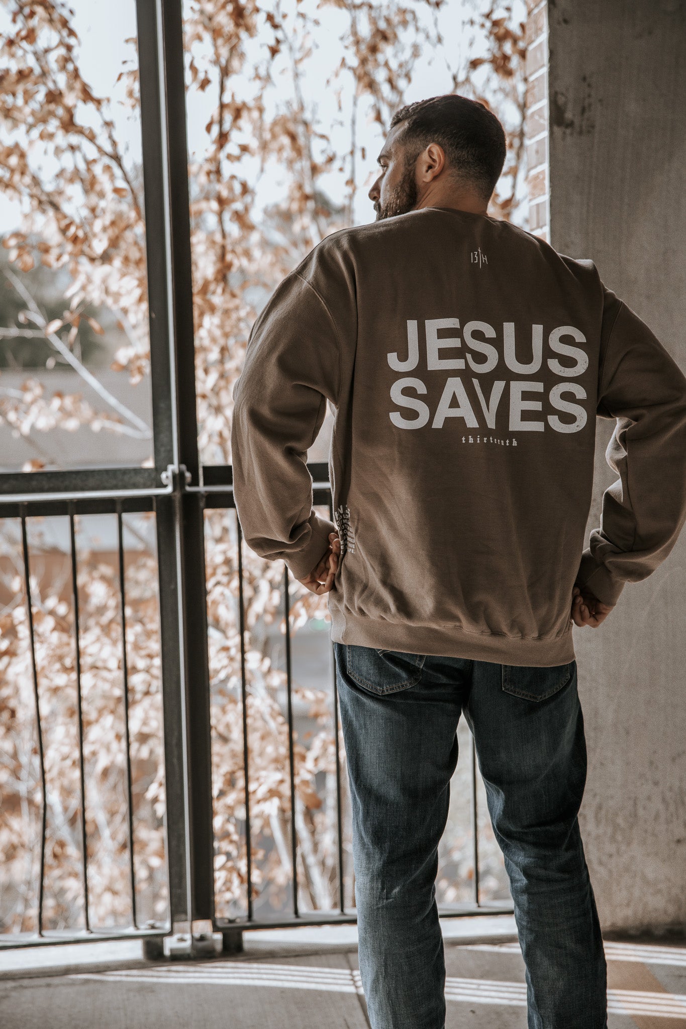 A man stands on a balcony in the 13th Jesus Saves Crewneck, featuring bold white "JESUS SAVES" text on the back. Trees with autumn leaves are visible behind him.