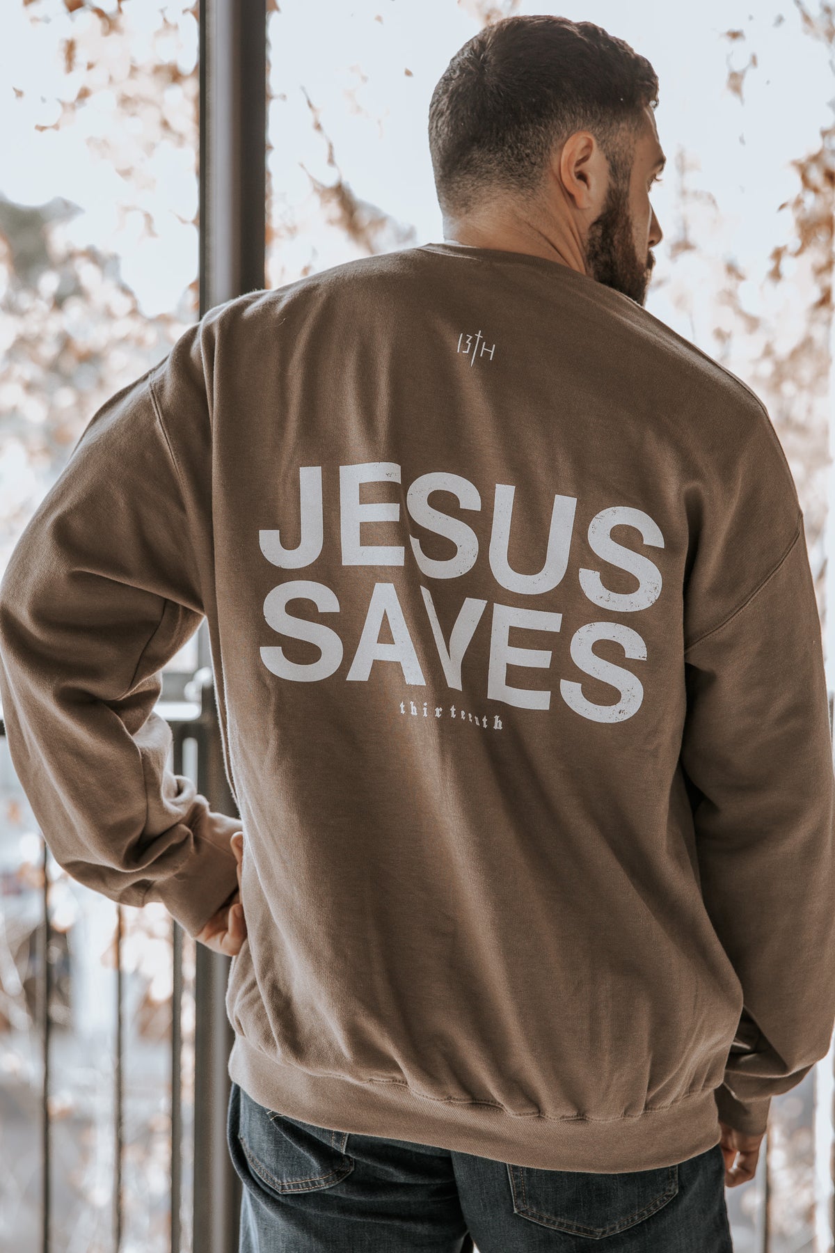 A man faces away from the camera in a 13th Jesus Saves Crewneck, featuring bold white "JESUS SAVES" text on the back. Trees and bright light appear in the background.