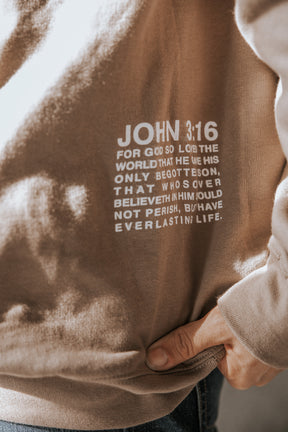 A close-up of the tan Jesus Saves Crewneck by 13th, featuring a hand on the hip and displaying "John 3:16 For God so loved the world..." on the front.