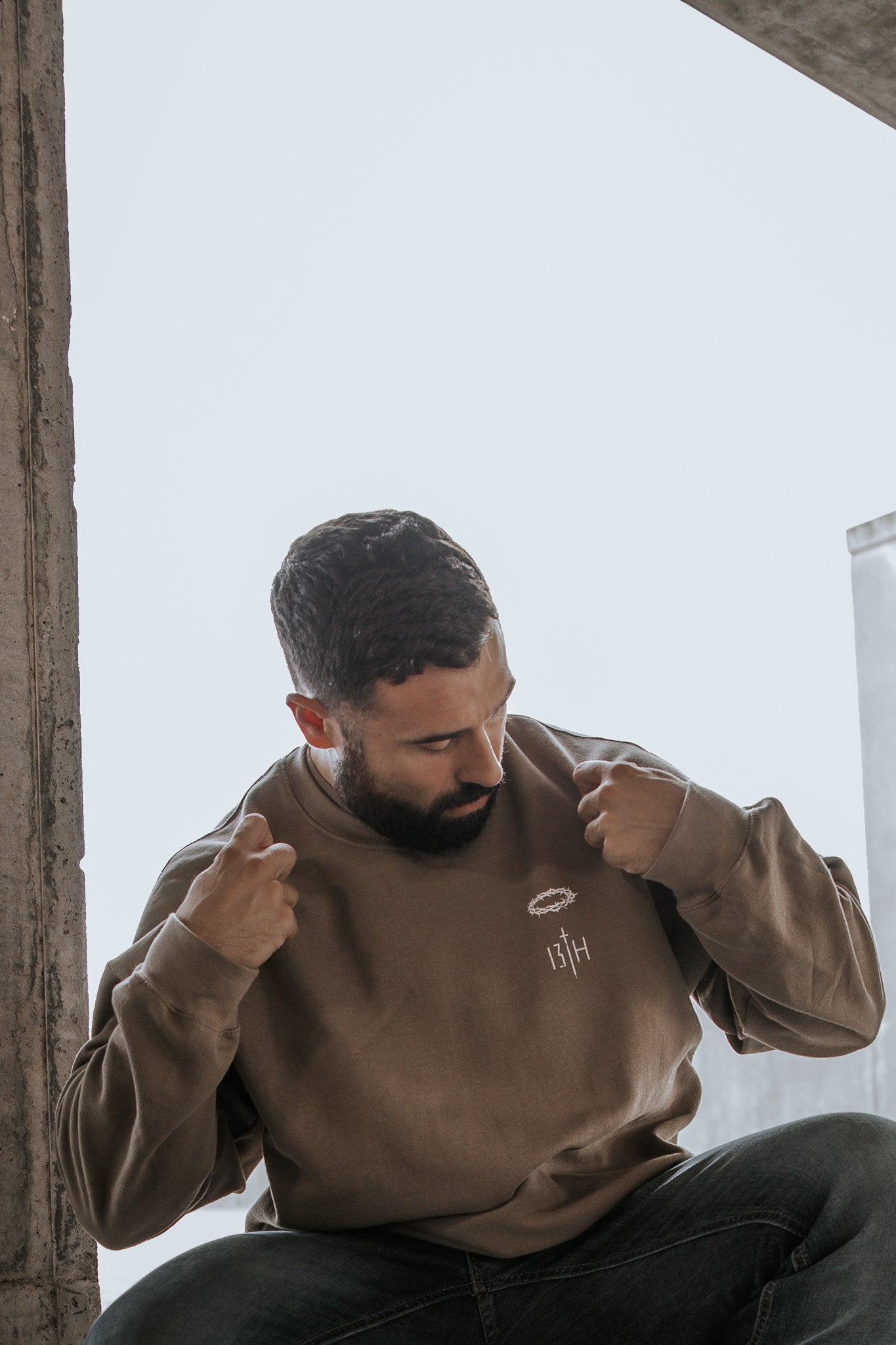 A bearded man with short hair sits indoors, looking down and pulling at the collar of his oversized 13th Jesus Saves Crewneck. He appears thoughtful against a softly lit, minimal background.