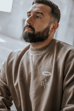A bearded man with short hair wears the 13th Jesus Saves Crewneck, a taupe sweatshirt with a small embroidered crown of thorns and the number 13TH, gazing thoughtfully into the distance.