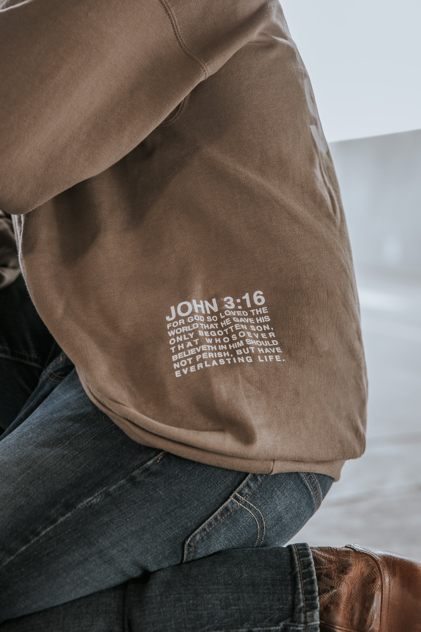 A person wearing the 13th Jesus Saves Crewneck, featuring John 3:16 in white text, paired with blue jeans and brown shoes, kneels on a light-colored floor.