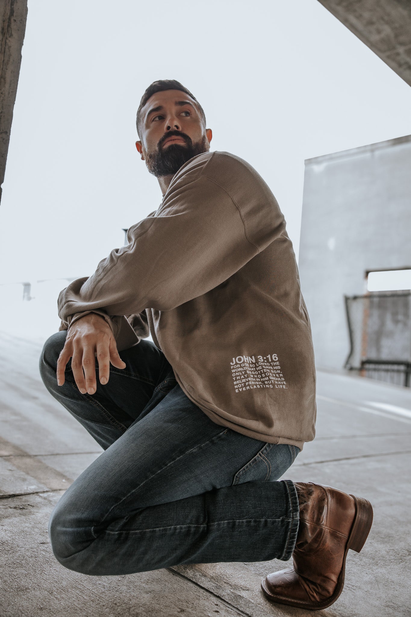 A bearded man wears the 13th Jesus Saves Crewneck, a tan long-sleeve shirt, paired with jeans and brown boots as he kneels in an outdoor urban setting, looking off to the side.