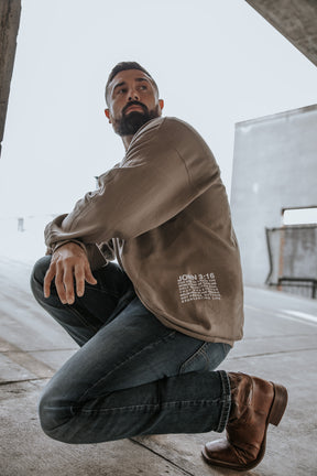 A bearded man wears the 13th Jesus Saves Crewneck, a tan long-sleeve shirt, paired with jeans and brown boots as he kneels in an outdoor urban setting, looking off to the side.
