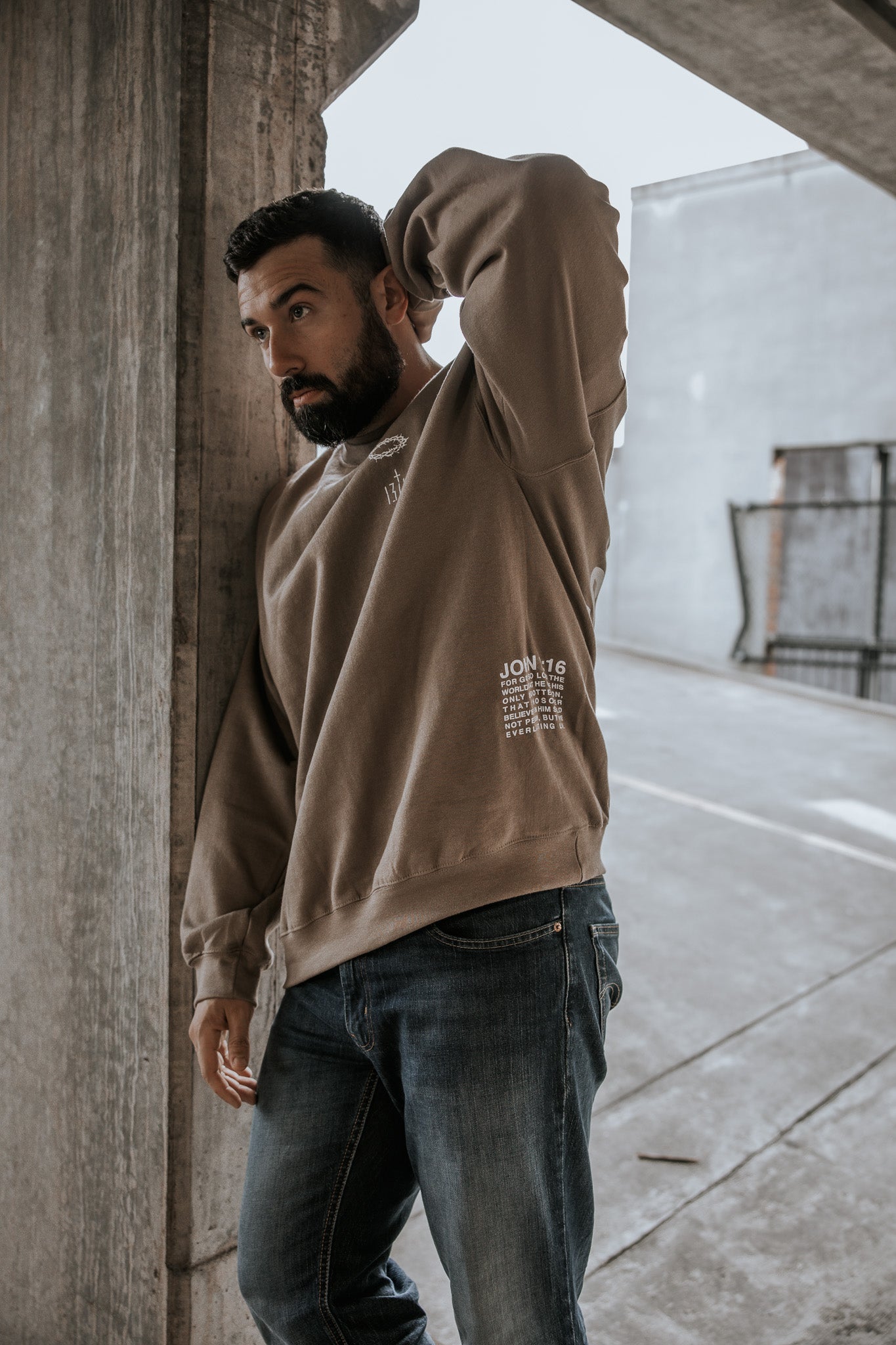 A bearded man stands by a concrete pillar in a parking garage, wearing the 13th "Jesus Saves" Crewneck and blue jeans, gazing thoughtfully into the distance with one hand behind his head.