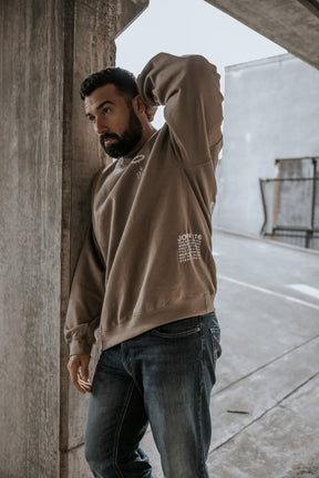 A bearded man stands by a concrete pillar in a parking garage, wearing the 13th "Jesus Saves" Crewneck and blue jeans, gazing thoughtfully into the distance with one hand behind his head.