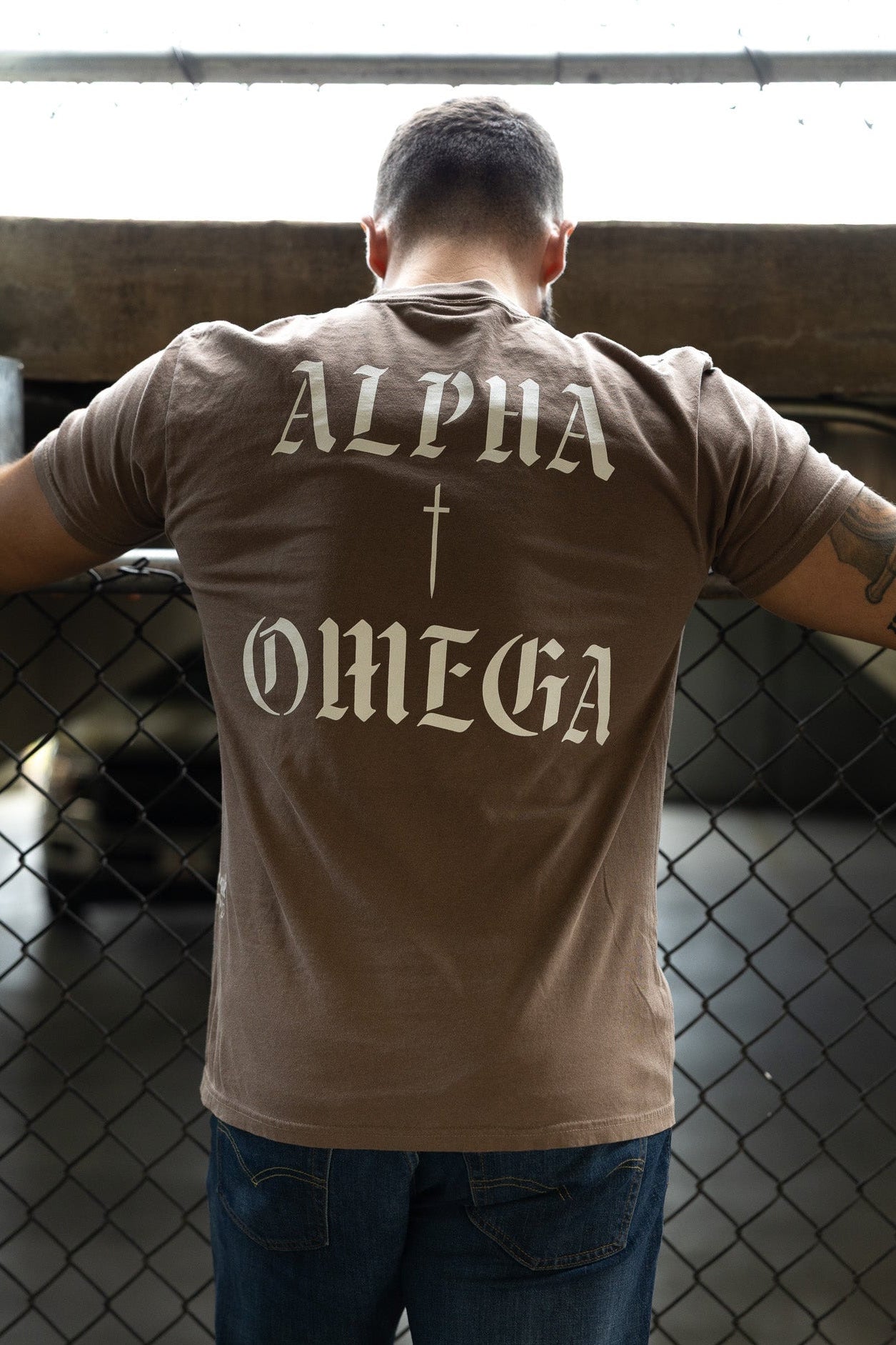 A man wearing the 13th Alpha & Omega Premium Oversized Tee in Espresso stands by a chain-link fence with his arms resting on the top rail in a dimly lit parking area.