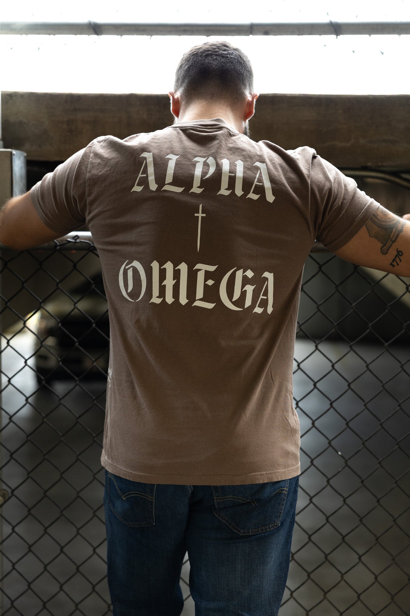 A man wearing the 13th Alpha & Omega Premium Oversized Tee in Espresso stands by a chain-link fence with his arms resting on the top rail in a dimly lit parking area.