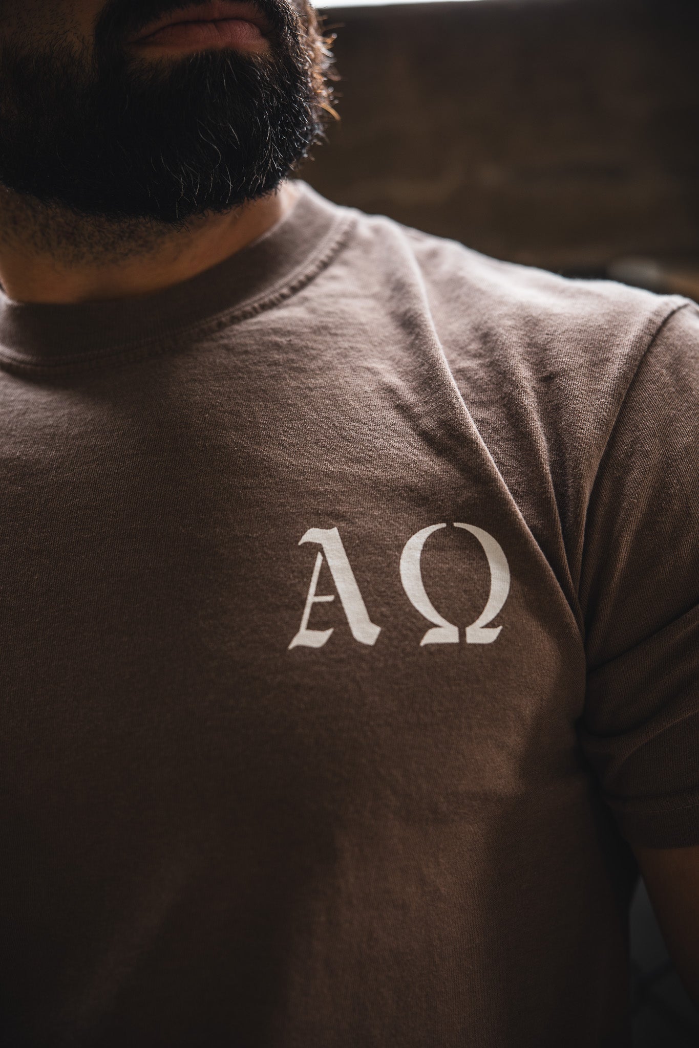 A bearded person wears the 13th Alpha & Omega Premium Oversized Tee in Espresso, featuring white Greek letters Alpha (Α) and Omega (Ω) on the chest.