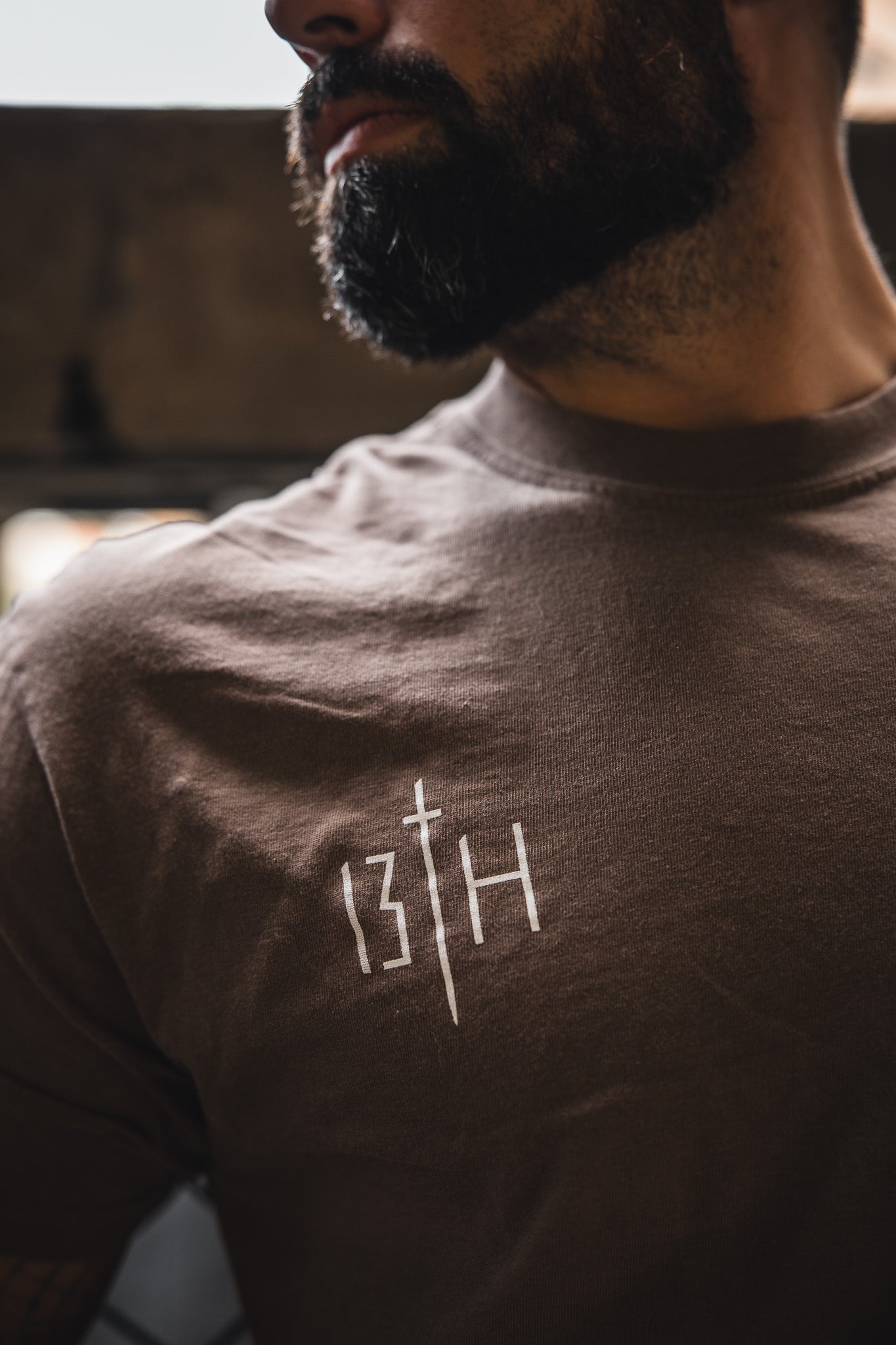 A bearded man wears the 13th Alpha & Omega Premium Oversized Tee in Espresso, featuring a white graphic with the number 13, a vertical sword, and the letter H; the background is softly blurred.