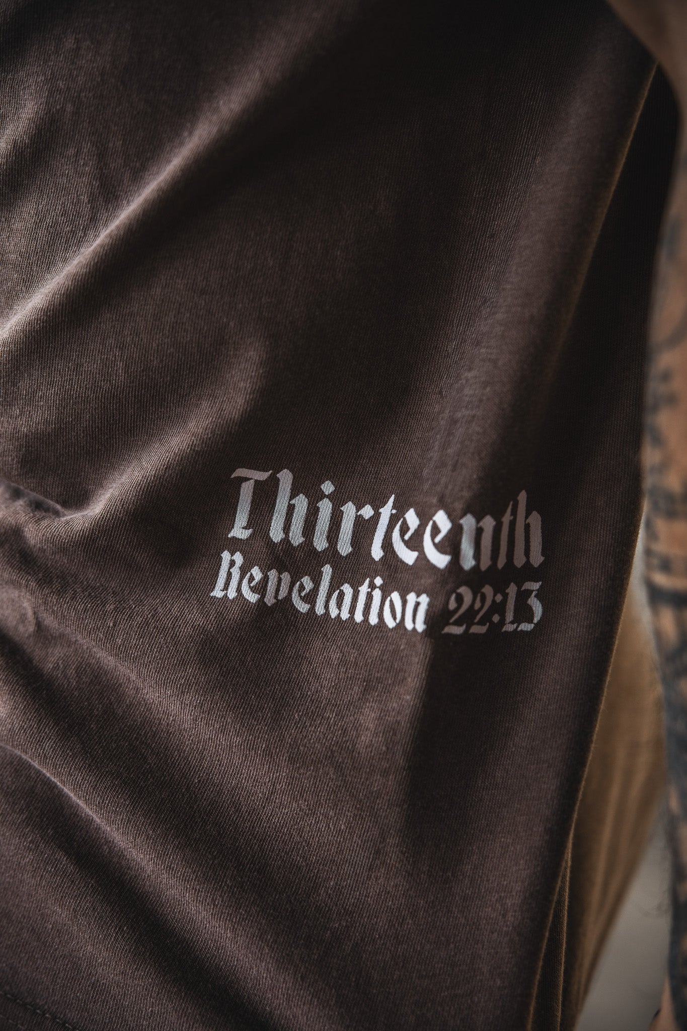 Close-up of the 13th Alpha & Omega Premium Oversized Tee in Espresso, showing brown fabric with white gothic-style “Thirteenth Revelation 22:13” text. A tattooed arm appears on the right.