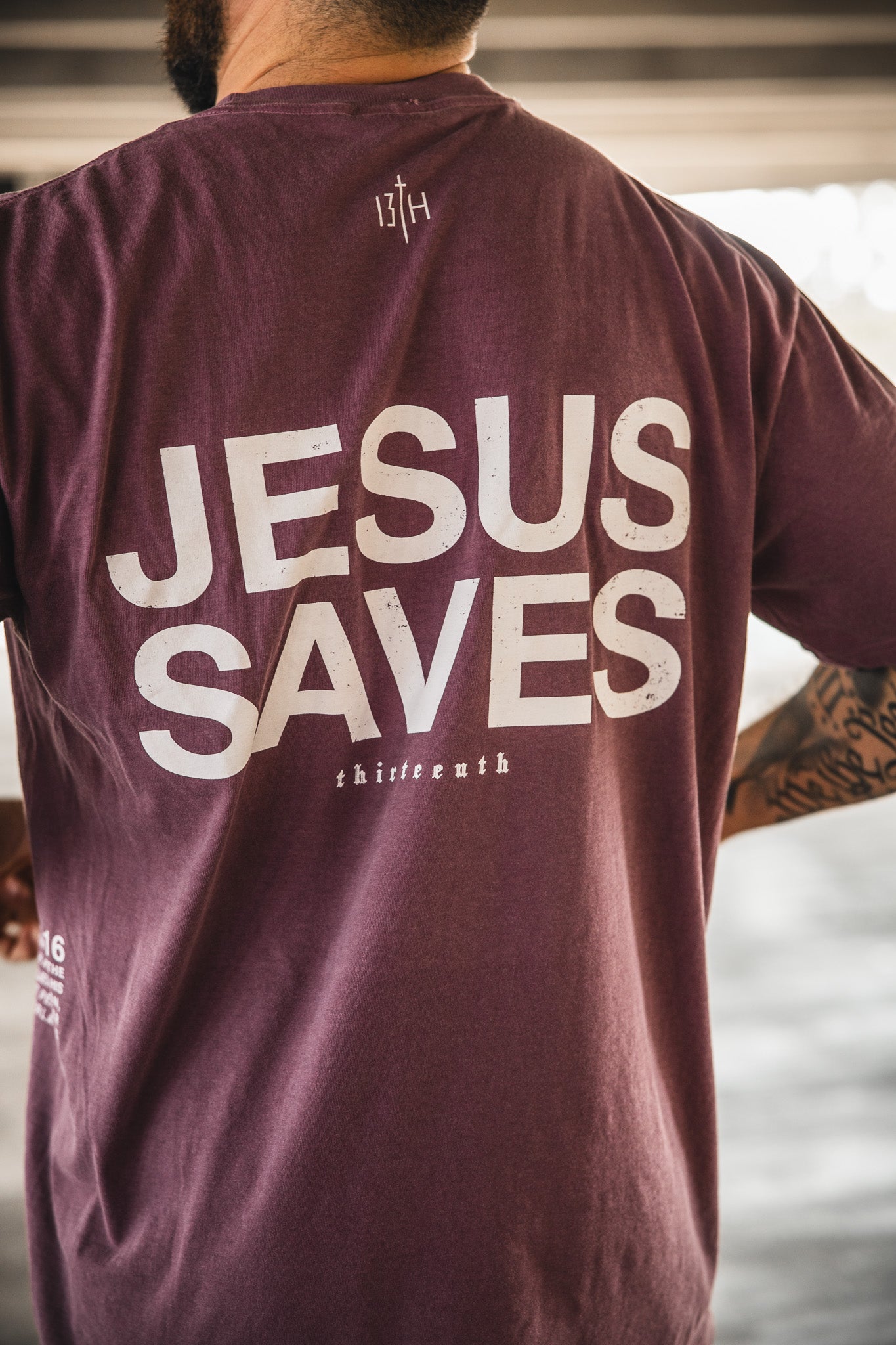 A person with tattoos on their right arm is standing indoors, wearing the 13th Jesus Saves Berry Tee. The maroon t-shirt features large white "JESUS SAVES" text printed on the back.