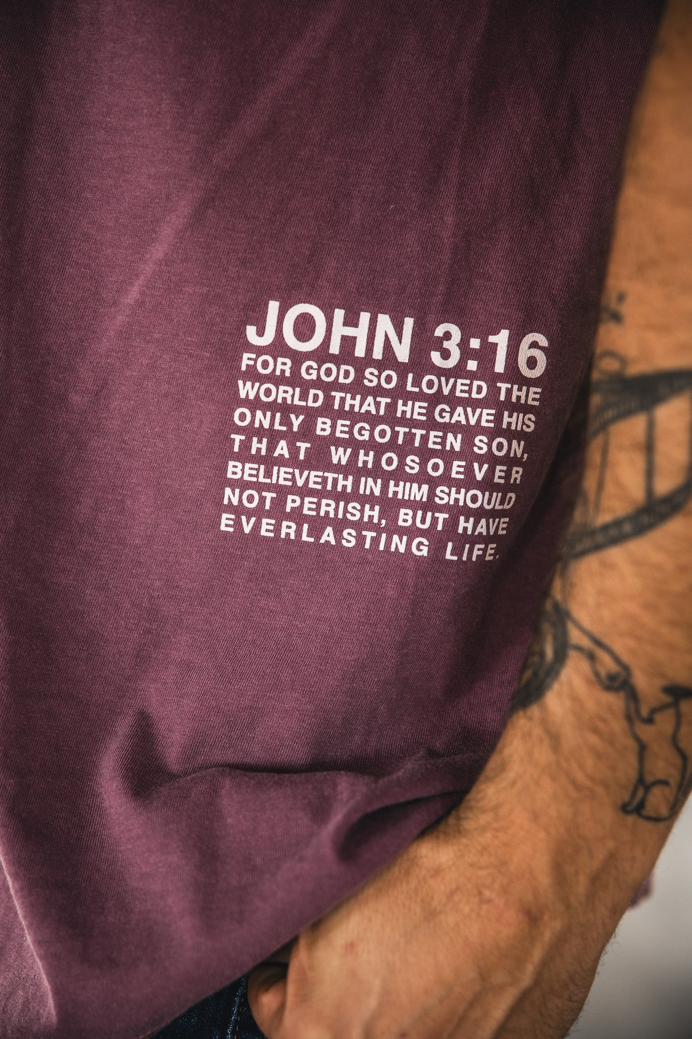 Close-up of the 13th Jesus Saves Berry Tee, featuring "John 3:16" in white text. Part of a tattooed arm appears beside the maroon shirt.