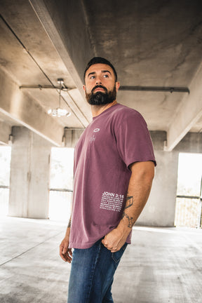 A bearded man stands in a sunlit, industrial-style parking garage wearing the 13th “Jesus Saves Berry Tee,” a mauve shirt with white John 3:16 text, paired with blue jeans, as he gazes off to the side.