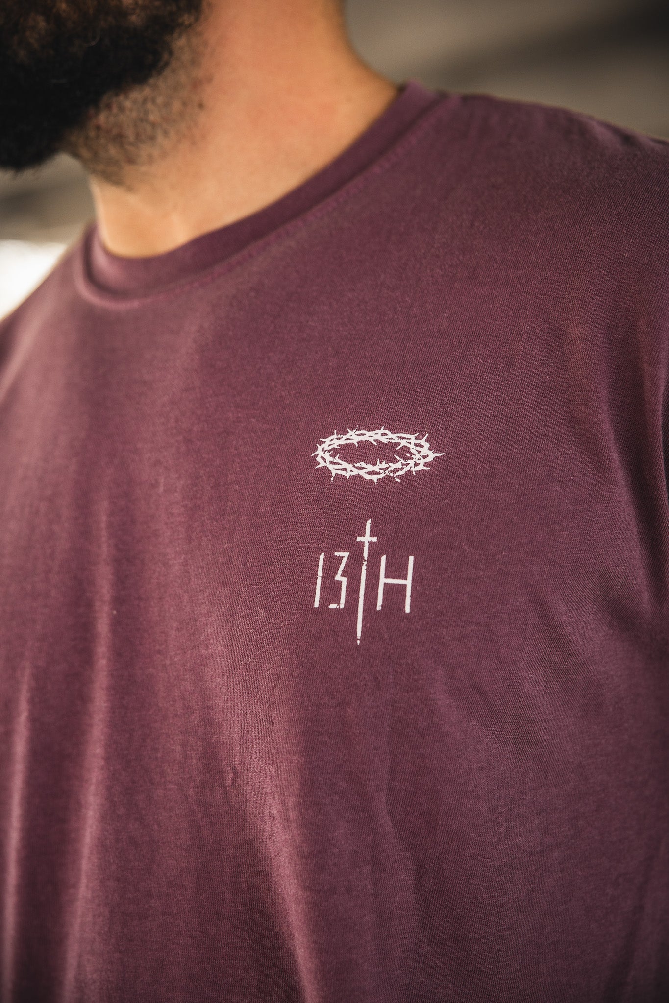 A close-up shows a person wearing the Jesus Saves Berry Tee by 13th, featuring a maroon color and a white crown of thorns graphic above "13TH" with the T as a cross. The person's face is partially out of frame.
