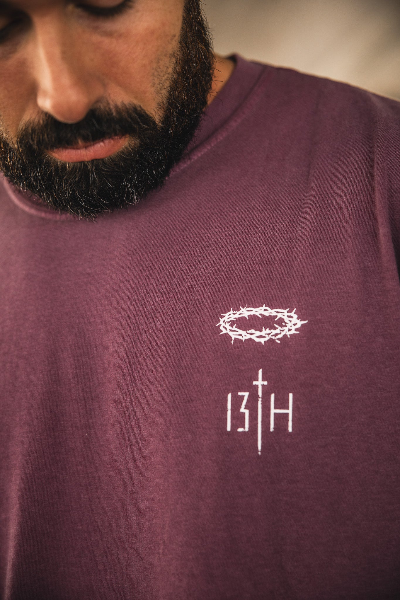 Close-up of a man wearing the 13th Jesus Saves Berry Tee, featuring a white crown of thorns graphic above "13TH" with the T styled as a cross. The man's face is partially visible, and he is dressed in maroon.