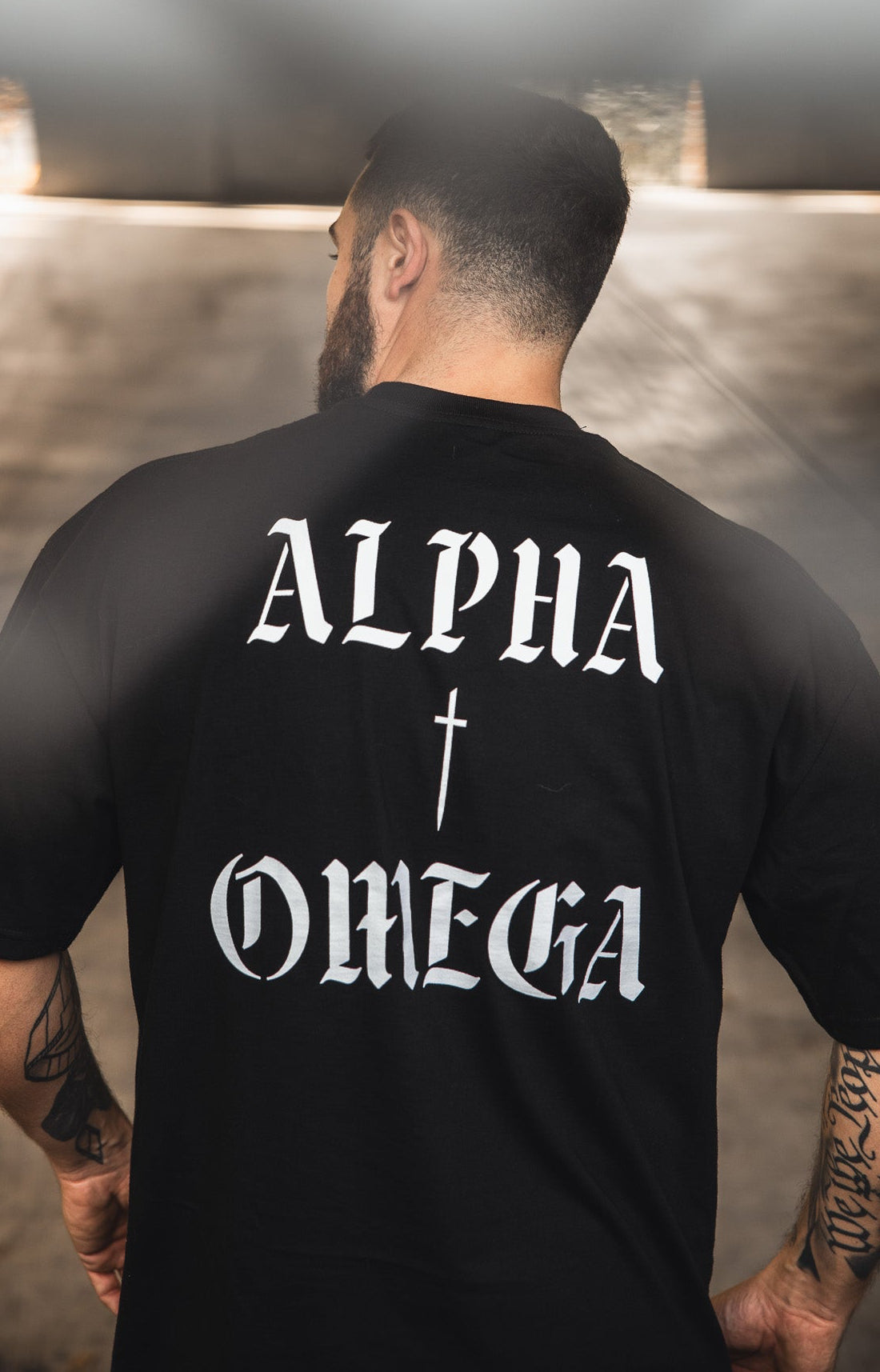 A man with short hair and tattoos faces away, wearing the 13th Alpha & Omega Premium Oversized Tee in black, featuring “ALPHA” and “OMEGA” in bold white letters with a cross symbol between them.