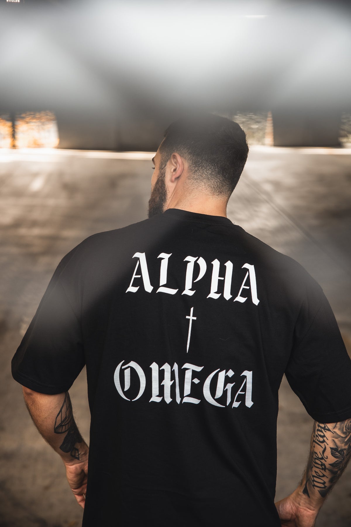 A man with short hair and tattoos faces away, wearing the 13th Alpha & Omega Premium Oversized Tee in black, featuring “ALPHA” and “OMEGA” in bold white letters with a cross symbol between them.