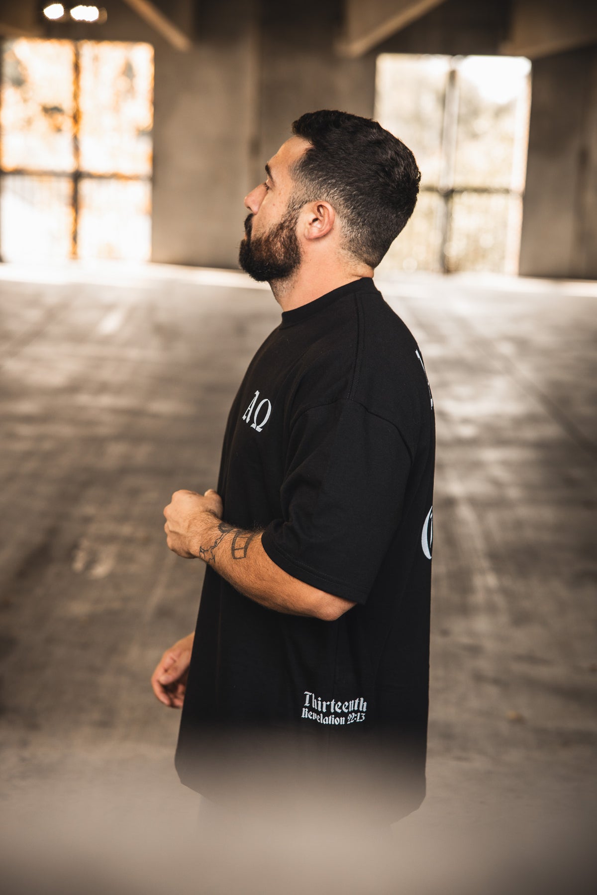 A man with a beard and short hair stands sideways in a sunlit concrete space, wearing the 13th Alpha & Omega Premium Oversized Tee in black with white side and back text, looking forward neutrally.