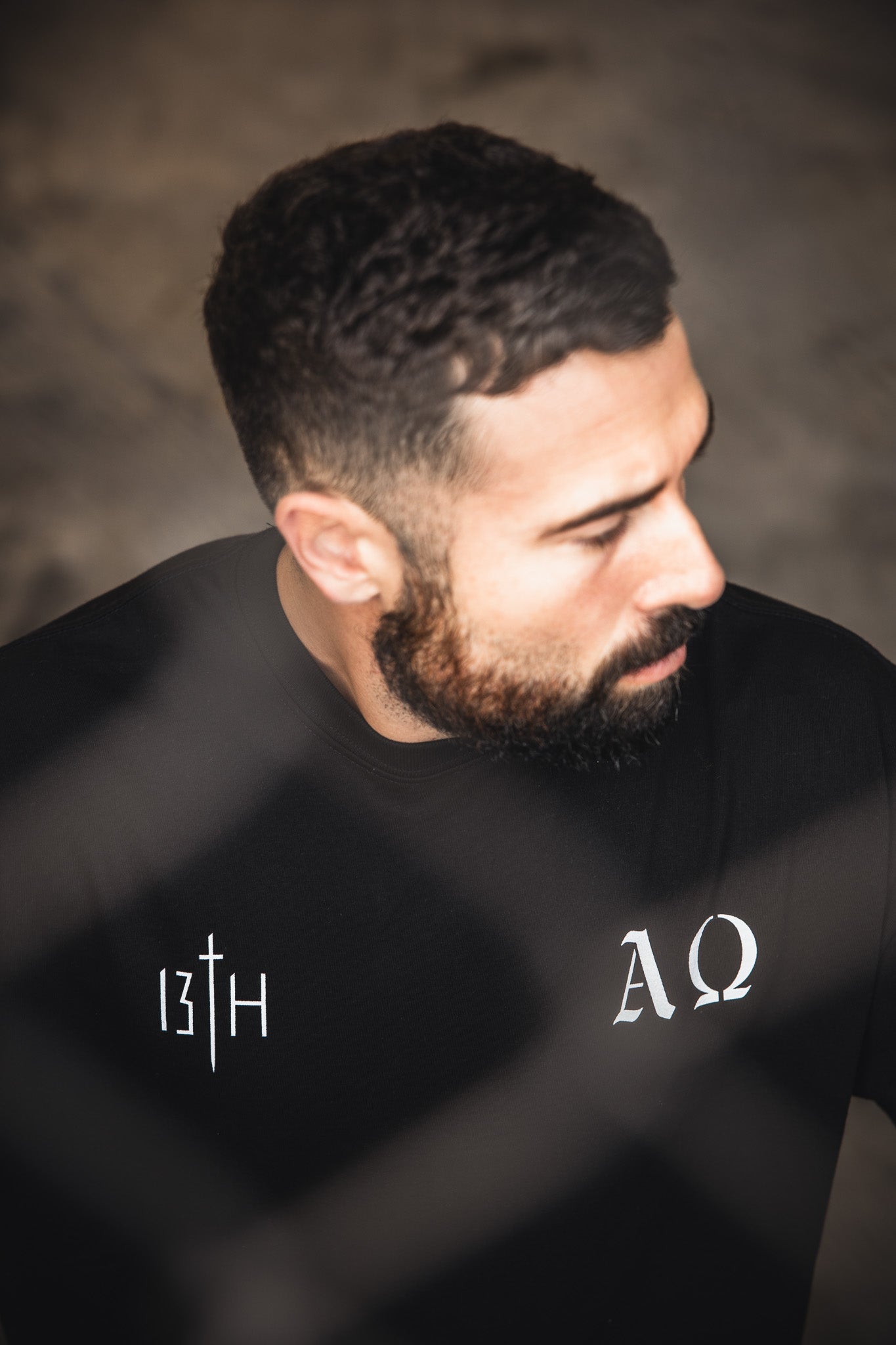 A bearded man with short dark hair looks down and away, wearing the 13th Alpha & Omega Premium Oversized Tee in black, featuring white "13th" and "AΩ" symbols on the chest, with a blurred wire fence in the foreground.
