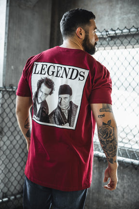 A tattooed man stands by a metal fence in an industrial area, wearing the Born United Private Label “Legends - Wet Bandits” red t-shirt with a black-and-white graphic of two men and the word LEGENDS on the back.