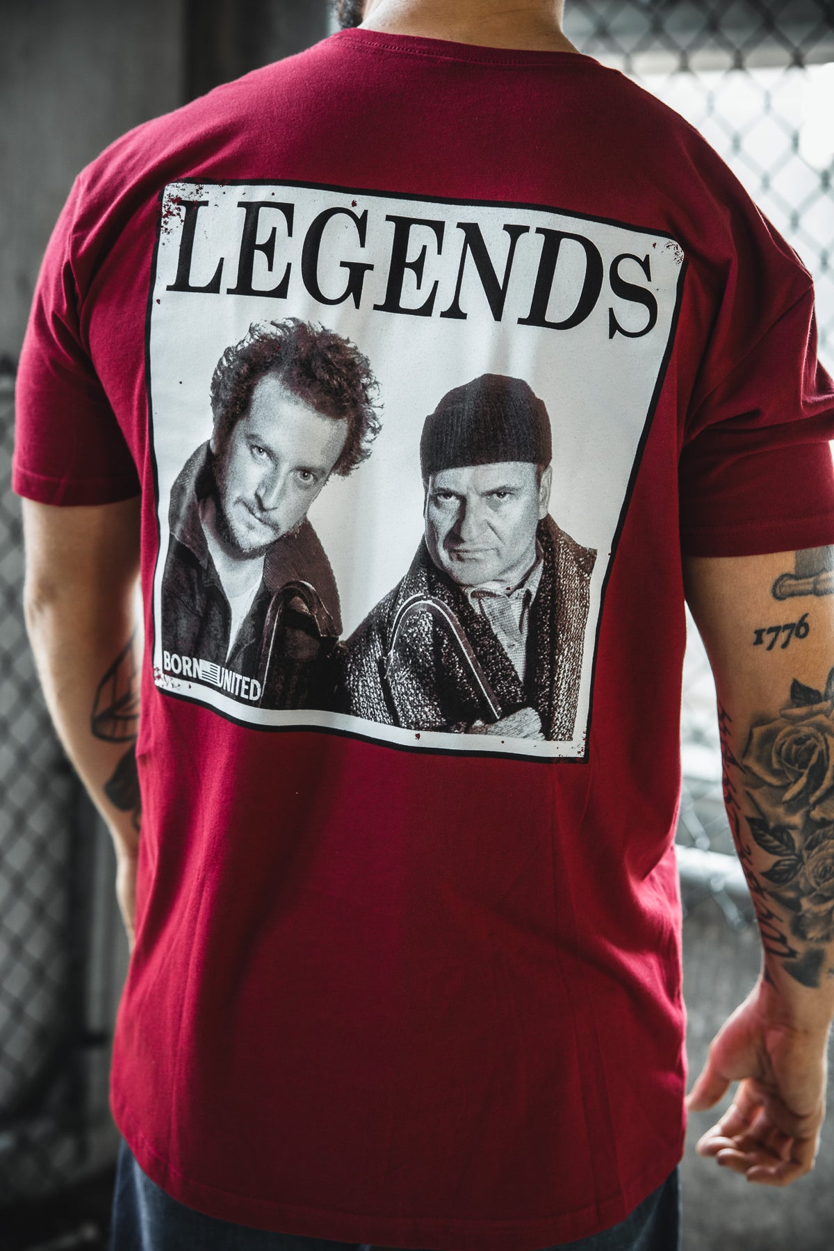 A person wears the Born United Private Label "Legends - Wet Bandits" red t-shirt with a black-and-white image of two men and the word LEGENDS on the back. The blurred background has a chain-link fence and lighting.