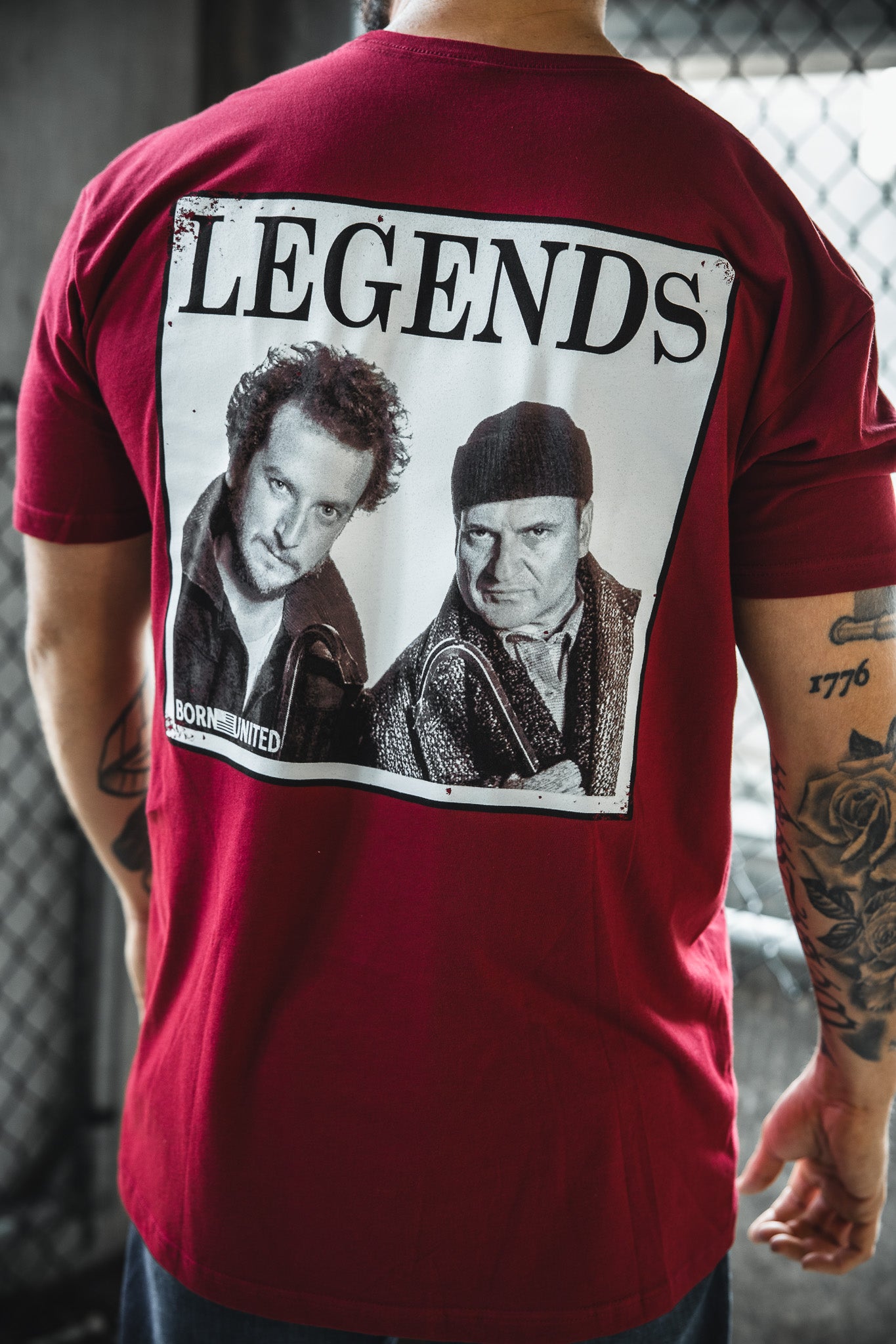 A person wears the Born United Private Label "Legends - Wet Bandits" red t-shirt with a black-and-white image of two men and the word LEGENDS on the back. The blurred background has a chain-link fence and lighting.