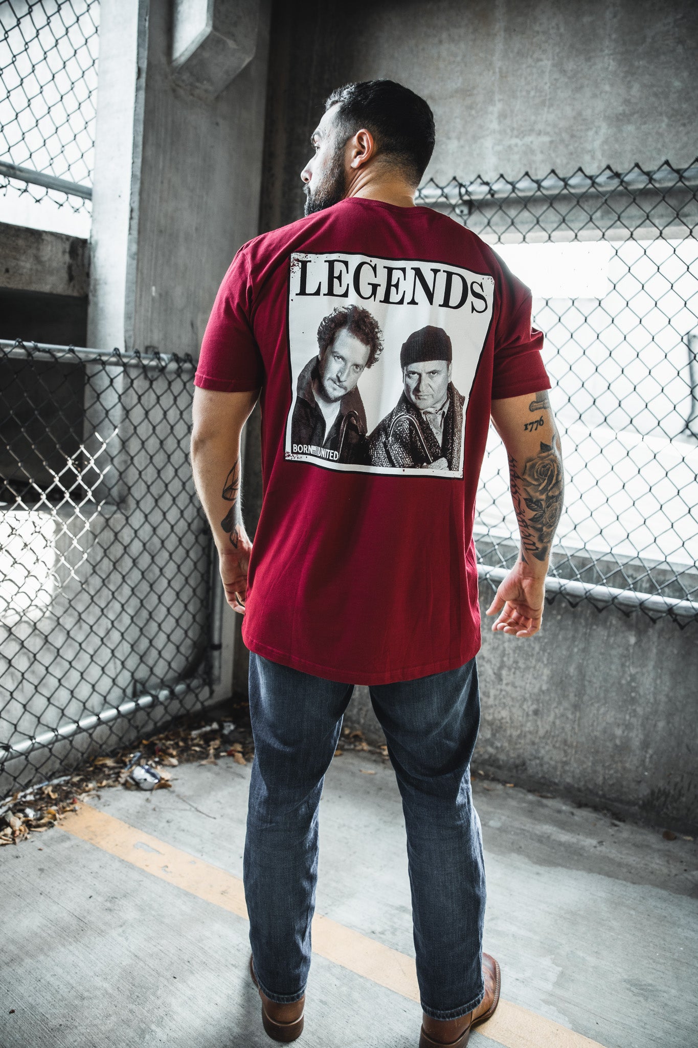 A man stands with his back to the camera, wearing the Born United Private Label "Legends - Wet Bandits" red T-shirt. He is in a concrete parking area next to a chain-link fence.