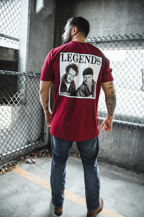 A man stands with his back to the camera, wearing the Born United Private Label "Legends - Wet Bandits" red T-shirt. He is in a concrete parking area next to a chain-link fence.