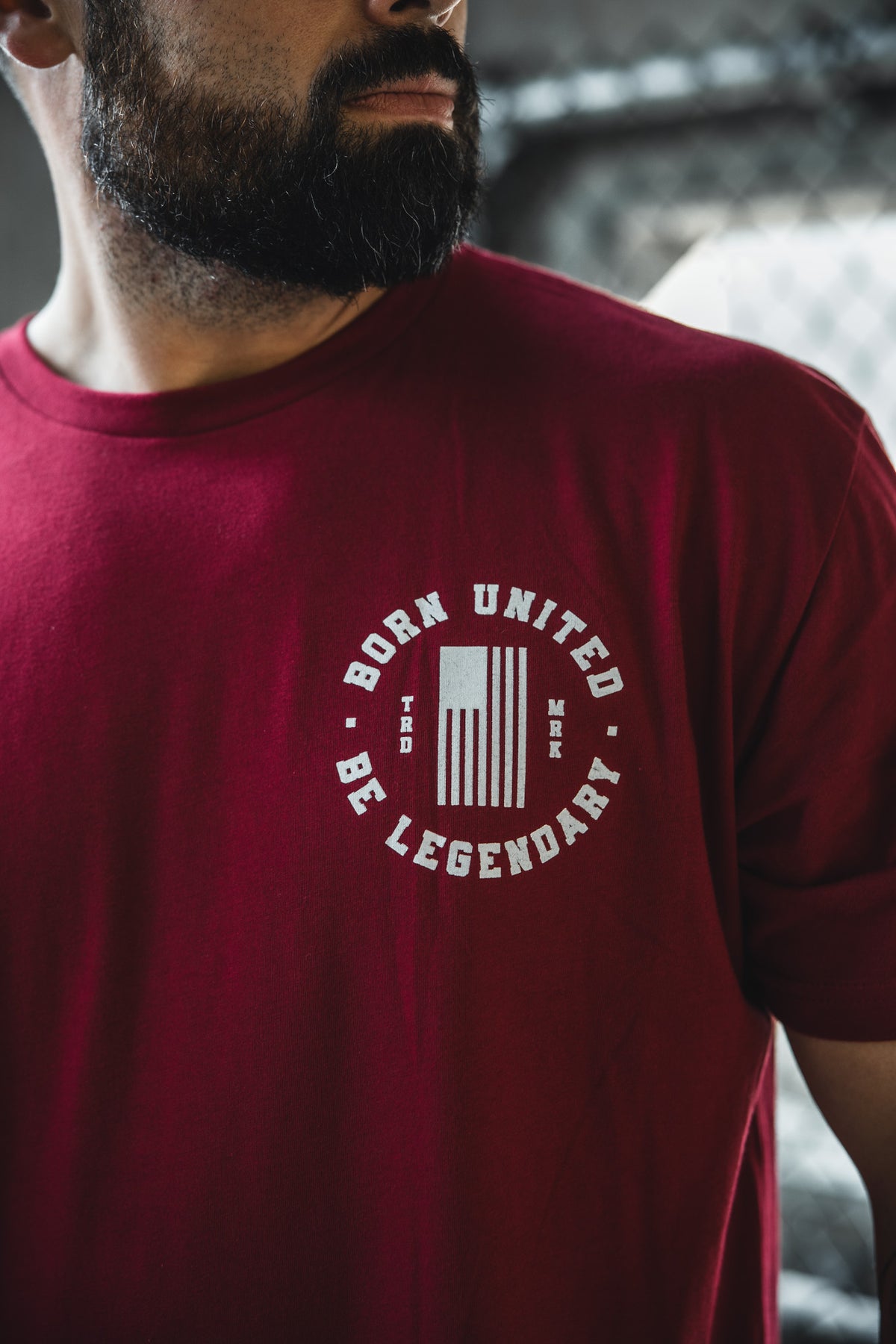 A bearded man wears a red Born United Private Label T-shirt featuring a circular white "BORN UNITED BE LEGENDARY" logo, a vertical graphic, and small "TDD MRR" text from the Legends - Wet Bandits collection.