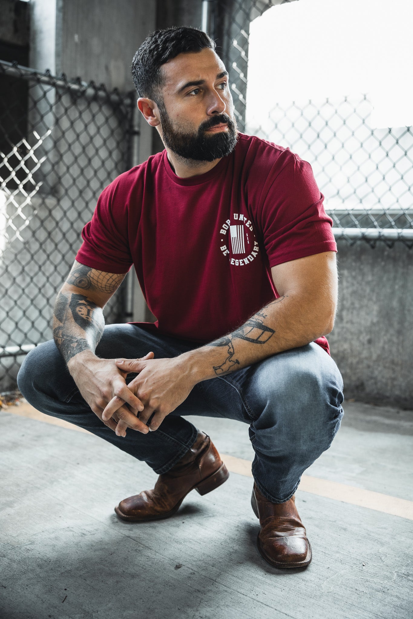 A bearded, tattooed man wearing the Legends - Wet Bandits t-shirt by Born United Private Label squats in an outdoor urban area near a chain-link fence, glancing to the side.