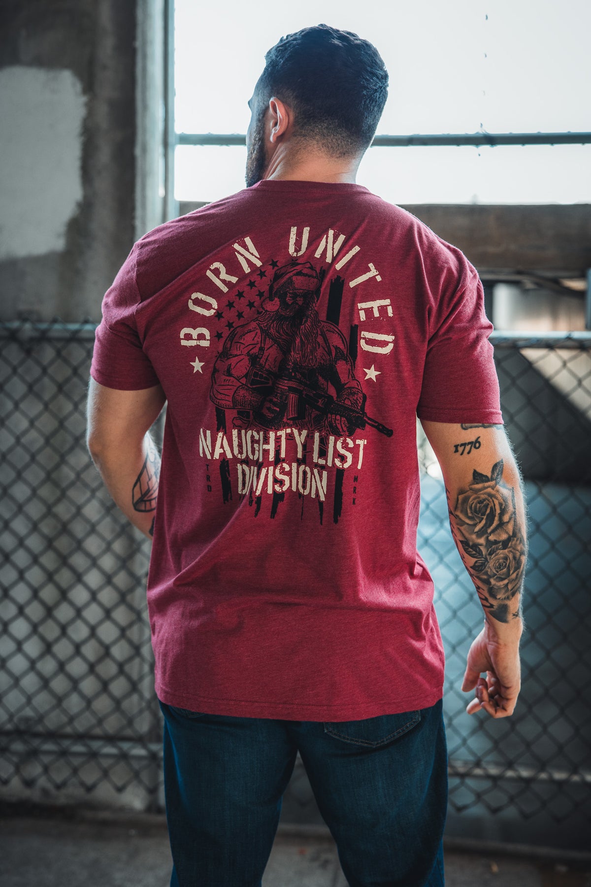 A tattooed man stands indoors by a chain-link fence, wearing the Born United Private Label Tactical Santa Tee—a limited-edition shirt featuring a soldier, American flag, and "BORN UNITED NAUGHTY LIST DIVISION" for bold Christmas spirit.